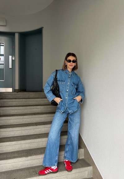 Conjunto de mezclilla con camisa de gran tamaño y jeans de pierna ancha en azul claro, combinado con zapatillas deportivas rojas con rayas blancas y un bolso negro texturizado.