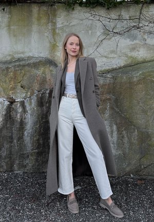 Mujer de pie al aire libre contra una pared de roca, vestida con un abrigo largo gris, pantalones blancos, camiseta blanca y mocasines grises.
