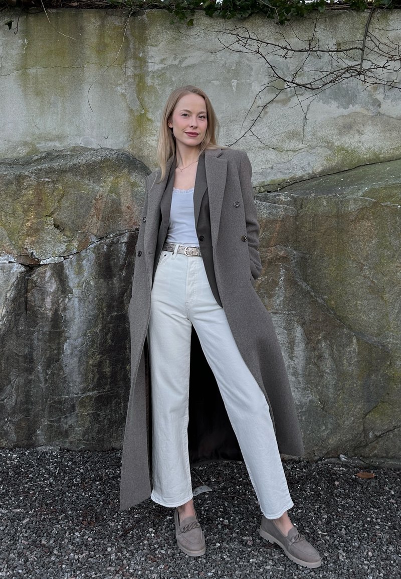 Femme debout en extérieur contre un mur de pierre, portant un long manteau gris, un pantalon blanc, un haut blanc et des mocassins gris.