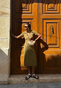 Olíva zöld, bordázott ruha rövid ujjakkal, slim fit fazonban, fonott táskával és ezüst szandállal párosítva, fából készült ajtó háttér előtt.