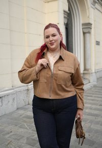 Bruine suède cropped jack met twee voorzakken, ritssluiting, gedragen over een kanttop. Donkere spijkerbroek en kleine bruine handtas.