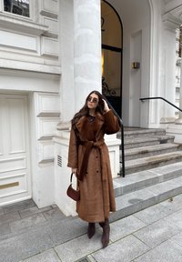 Brown textured long coat with a belt, double-breasted design, paired with dark sunglasses and a brown handbag, worn with brown pointed heels.