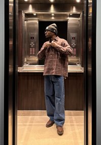 Brūna pledēta pogājama krekls virs melnas krekla, zili džinsi un brūnas čības, pelēka cepure, stāv metāla liftā.