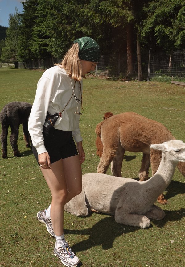 Eine junge Person in einem weißen Langarmshirt und schwarzen Shorts steht auf grünem Gras neben hellen und dunklen Alpakas. Ein grünes Bandana und Sneakers sind sichtbar.