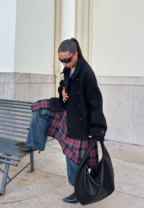 Une femme portant des lunettes de soleil et un manteau noir pose à côté d’un banc en métal. Elle porte une jupe à carreaux et un jean, et tient un grand sac noir.