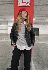 Μαύρο δερμάτινο μπουφάν μηχανής, γκρι πουλόβερ και λευκό πουκάμισο layered από κάτω. Κόκκινη μπερέ, μαύρο παντελόνι, ποζάροντας μπροστά σε έναν κόκκινο τοίχο σε μια περιοχή στάθμευσης.