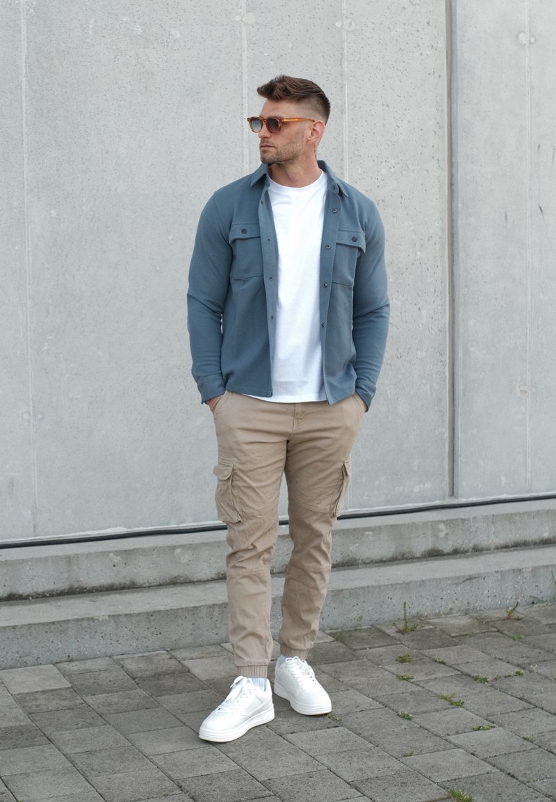 Homme portant des lunettes de soleil, une surchemise bleue, un t-shirt blanc, un pantalon cargo beige et des baskets blanches, debout contre un mur en béton gris.