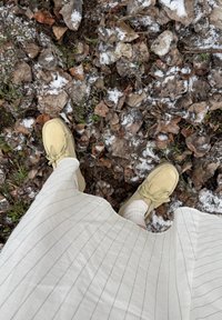 Pieds en chaussures beige et chaussettes blanches debout sur un sol couvert de feuilles sèches et de neige légère, portant une jupe blanche à fines rayures.