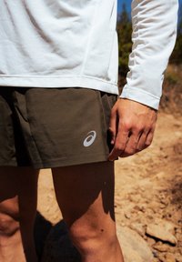 Calções castanhos curtos com um logótipo prateado na perna, combinados com uma camisa de mangas longas branca texturizada. Os detalhes incluem costura visível e um ambiente exterior.