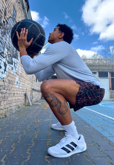 Hombre en cuclillas al aire libre sosteniendo una pelota medicinal negra cerca de una pared de ladrillo cubierta de grafiti en una pista deportiva azul bajo un cielo parcialmente nublado.