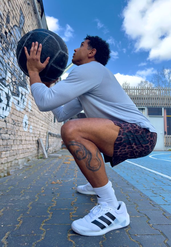 Άνδρας κάθισμένος σε καθίσμα έξω, κρατώντας μια μαύρη ιατρική μπάλα κοντά σε έναν τοίχο από τούβλα με γκράφιτι, σε γήπεδο μπλε αθλητισμού κάτω από ένα μερικώς συννεφιασμένο ουρανό.