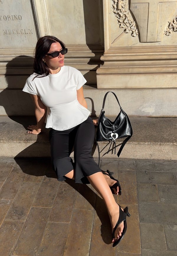Une femme portant des lunettes de soleil est assise au soleil, à côté d’un sac à main noir. Elle porte un haut blanc, un pantalon noir et des sandales, devant un mur de pierre sculpté.
