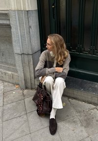Siva i bež džemper s geometrijskim uzorkom, kremska košulja s ovratnikom, bijele hlače, tamnosmeđa torba s maramom s točkastim uzorkom; sjedi na gradskom pločniku.