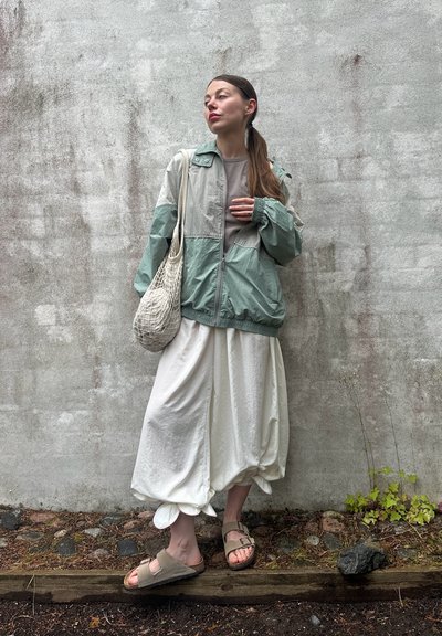 Chaqueta ligera en verde menta y gris con puños elásticos, combinada con pantalones blancos holgados y sandalias grises. Una bolsa de red texturizada acentúa el conjunto.