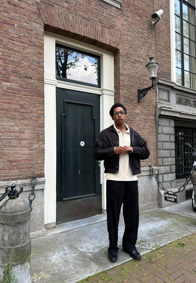 Chaqueta negra de cremallera sobre una polo beige, pantalones anchos negros y zapatos negros. Muro de ladrillo y puerta oscura al fondo.