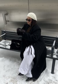 Femme en béret blanc, manteau noir et gants, assise sur un banc poudré de neige, ajustant ses lunettes de soleil, avec un sac à main noir à côté d'elle.