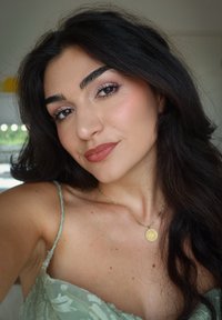 Persona che indossa un abito floreale verde chiaro con spalline sottili, una collana d'oro con un pendente tondo e un trucco naturale con toni delicati.