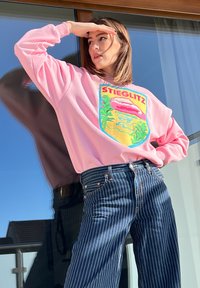 Sweatshirt rosa claro com um gráfico colorido contendo o texto "STIEGLITZ" e um design natural, combinado com jeans de denim listrados em azul e branco.
