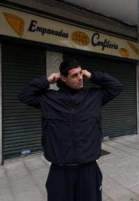 Rompevientos negro con cierre, de corte suelto y tejido texturizado, combinado con pantalones de chándal oscuros que tienen un pequeño logo. El fondo incluye carteles de la tienda.