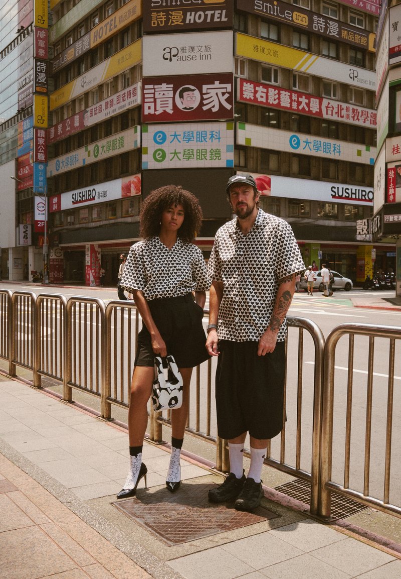 Duas pessoas estão na rua de uma cidade, vestindo camisas de manga curta com padrões preto e branco combinando. A mulher usa calções pretos e uma bolsa estampada. O homem veste calções pretos, ténis e um boné. O fundo apresenta letreiros coloridos de lojas.