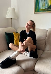 Čierne papuče s bielym lemovaním, útulné biele ponožky a čierny kardigán na béžovej pohovke so zelenými a žltými vankúšmi a lampou.