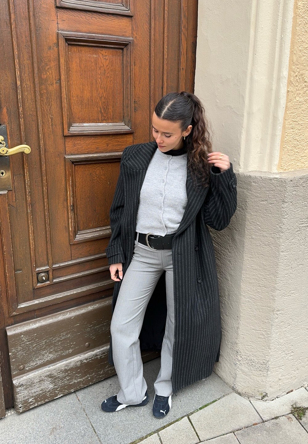 Lungo cappotto nero a righe, maglione grigio a maglia, pantaloni grigi svasati, cintura nera, scarpe da ginnastica, in piedi accanto a una porta di legno, guardando in basso.
