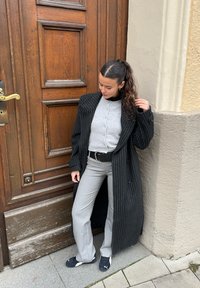 Lungo cappotto nero a righe, maglione grigio a maglia, pantaloni grigi svasati, cintura nera, scarpe da ginnastica, in piedi accanto a una porta di legno, guardando in basso.