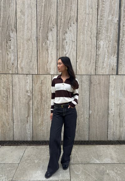 Camisa de rayas de manga larga en blanco y marrón oscuro, combinada con pantalones vaqueros de pierna ancha oscuros; de pie sobre una superficie de piedra frente a una pared texturizada.