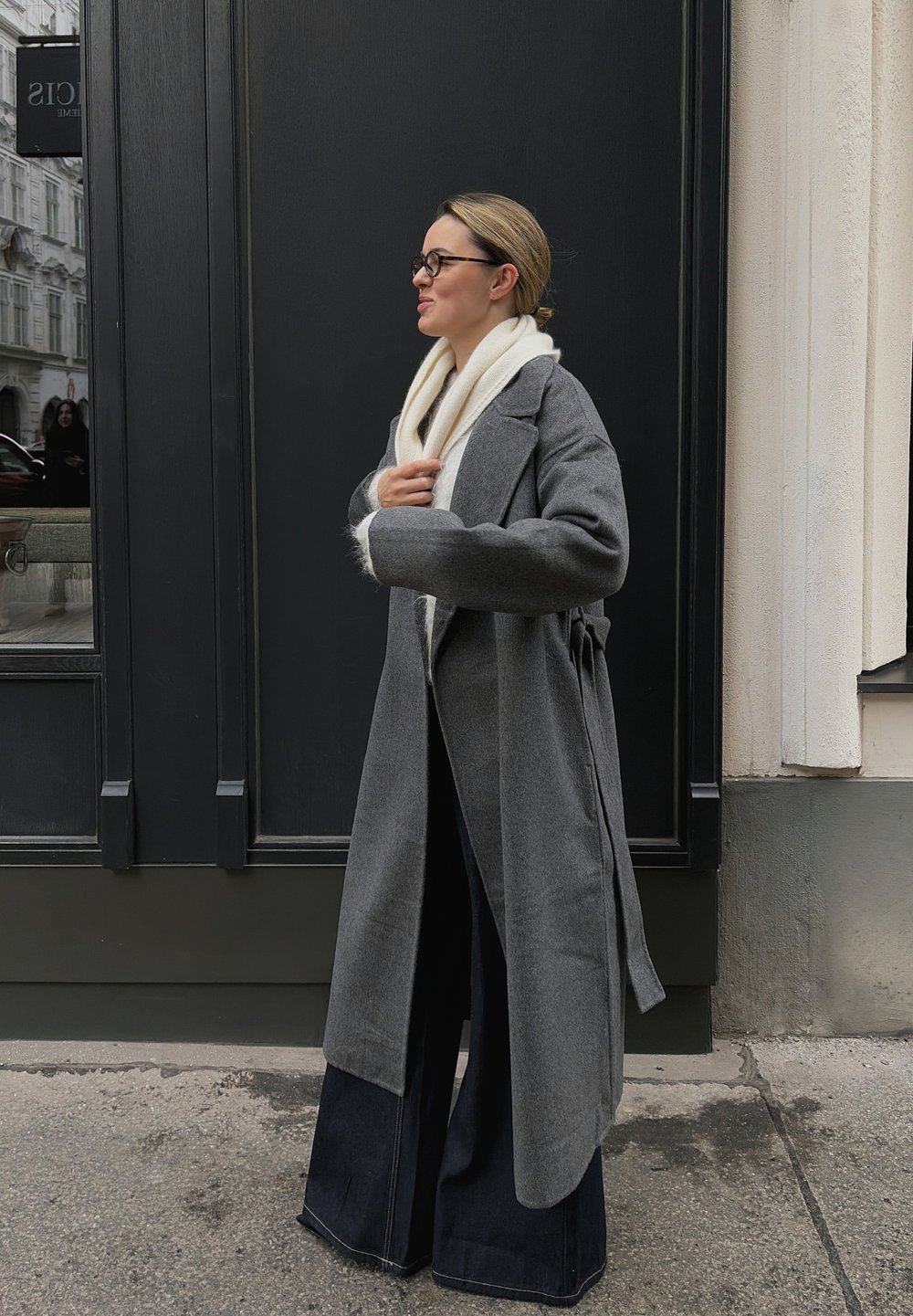 Lang grå kåpe med vide ermer, belte i midjen og et mykt hvitt skjerf. Modellen har på seg vide mørkeblå denimbukser. Bakgrunnen har en mørk vegg.