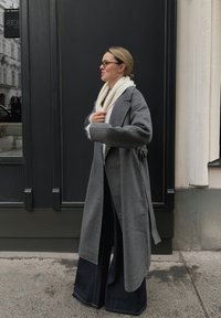 Lang grå kåpe med vide ermer, belte i midjen og et mykt hvitt skjerf. Modellen har på seg vide mørkeblå denimbukser. Bakgrunnen har en mørk vegg.