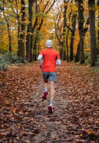 Yhdysvaltalaisessa metsässä juoksee juoksija kirkkaassa oranssissa paidassa ja harmaissa shortseissa, kun hän kulkee lehtien peittämällä polulla, jossa on keltaisia ja oransseja syksyn lehtiä.