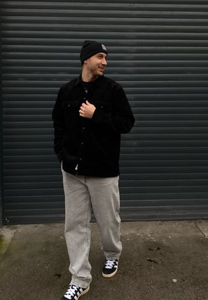 Chaqueta de pana negra con bolsillos frontales, jeans gris holgados y zapatillas negras y blancas, en un fondo texturizado gris.