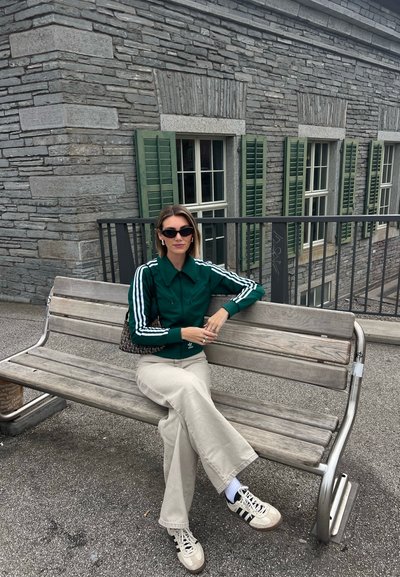 Chaqueta verde con cremallera y rayas blancas, pantalones de pierna ancha beige y zapatillas blancas. Sentado en un banco de madera contra una pared de piedra.