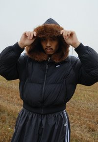 Doudoune noire avec capuche bordée de fourrure, manches longues et fermeture éclair à l'avant, associée à un pantalon de survêtement noir avec des bandes blanches.