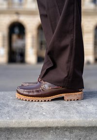 Braune Lederschuhe im Mokassin-Design, mit auffälligen Nähten und einer dicken, strukturierten Gummisohle. Kombiniert mit dunkelbraunen Hosen.