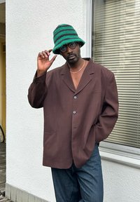 Blazer castanho oversized com dois botões e um bolso com aba, combinado com jeans escuros. Um chapéu de croché em azul e preto com riscas completa o visual.