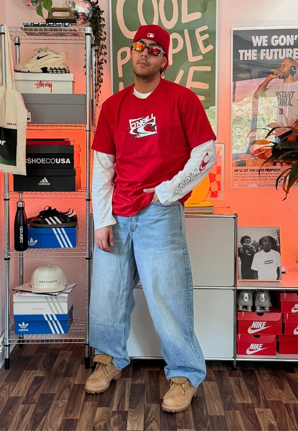 Man wearing red cap, red O'Neill shirt over white long sleeves, light blue baggy jeans, and tan boots in a room with shoe boxes and posters.