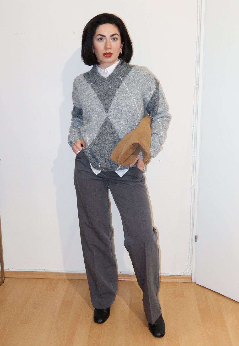 Femme portant un pull gris à losanges, une chemise blanche, un pantalon gris, des chaussures noires, tenant un chapeau beige, debout sur un sol en bois contre un mur blanc.