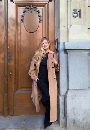 Beige Woll-Trenchcoat mit schwarzen Knöpfen, getragen über einem schwarzen Oberteil und einer Hose. Steht vor einer Holztür mit dekorativen Akzenten.