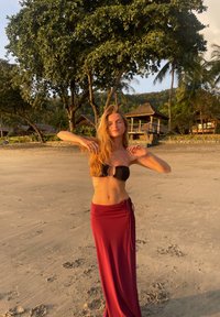 Černý bandeau bikinový top a dlouhá vínově červená zavinovací sukně. Hladká látka, uvázaná v pase, na písečné pláži s stromy v pozadí.