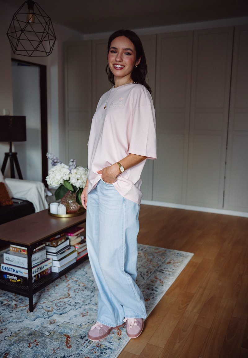 Jeune femme souriante, portant une chemise rose clair et un jean large bleu clair, debout dans un salon avec une décoration moderne et un tapis à motif.