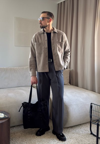 Hombre de pie en el interior, vistiendo chaqueta beige, camisa negra, pantalones grises, gafas de sol, sosteniendo una bolsa negra cerca de un sofá beige y cortinas.