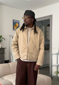 Bēša džemperis ar rāvējslēdzēju un apkakli, kombinēts ar kr�ēmkrāsas kreklu un brūnām biksēm. Džemperim ir neliels logotips sānos un zelta metāla detaļas.