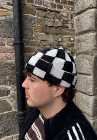Jeune homme portant un bonnet en tricot à carreaux noir et blanc et une veste sombre, debout contre un mur de pierre avec un tuyau noir, regardant vers la gauche de profil.