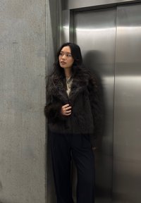 Jachetă din blană artificială maronie cu guler larg, purtată peste un top de culoare deschisă. Pantaloni largi, negri, și un fundal din oțel inoxidabil neted.