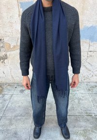 Homme portant un pull en maille gris foncé, une écharpe bleu marine avec des franges, un jean large délavé et des chaussures noires, debout sur un pavé texturé.