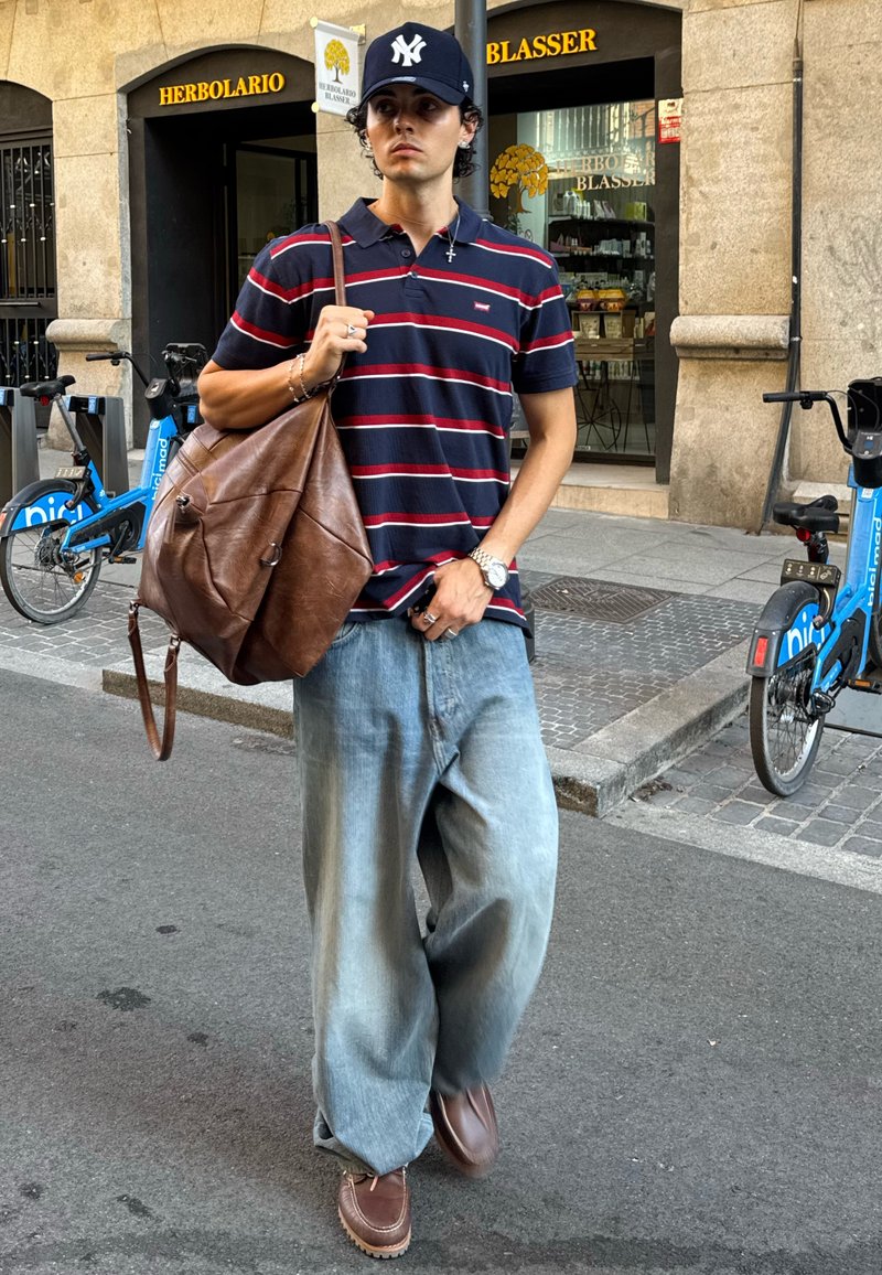 Jovem com polo às riscas e jeans largos, carregando uma grande bolsa de couro castanho, a caminhar numa rua da cidade perto de alugueres de bicicletas e da entrada de uma loja.