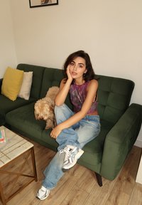 Sofá verde estofado com duas almofadas amarelas e brancas, chão de madeira clara, mesa de café com tampo de mármore, indivíduo com uma blusa roxa e jeans de ganga.