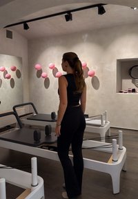 Pilates-reformer med en elegant hvid ramme og gråt polstret overflade, udstyret med sorte modstandsbånd og to sorte skulderstøtter. Pink dekor i baggrunden.