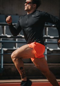 Mann in schwarzem Langarmshirt und orangefarbenen Shorts, der auf einer Außenbahn sprintet, im Hintergrund leere Zuschauertribünen.